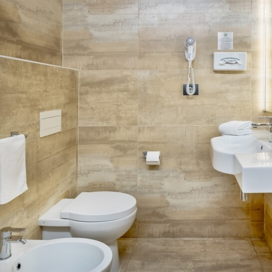 Bagno minimalista con toilet, bidet, lavabo e accessori, pareti beige e luce naturale da una finestra.