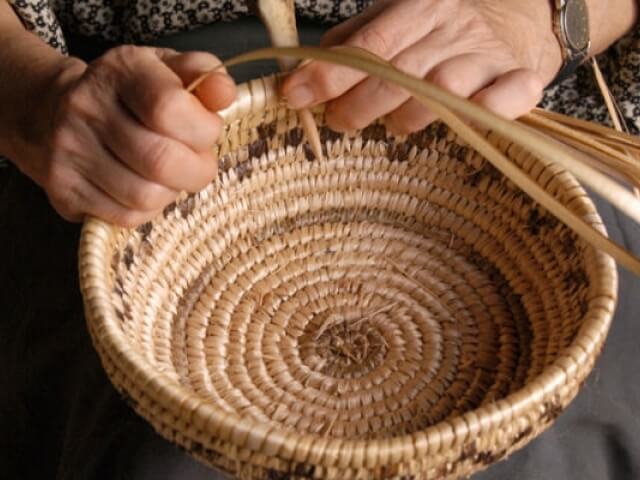 Le mani di una persona intrecciano un cesto circolare di giunco chiaro, con un motivo a spirale al centro.