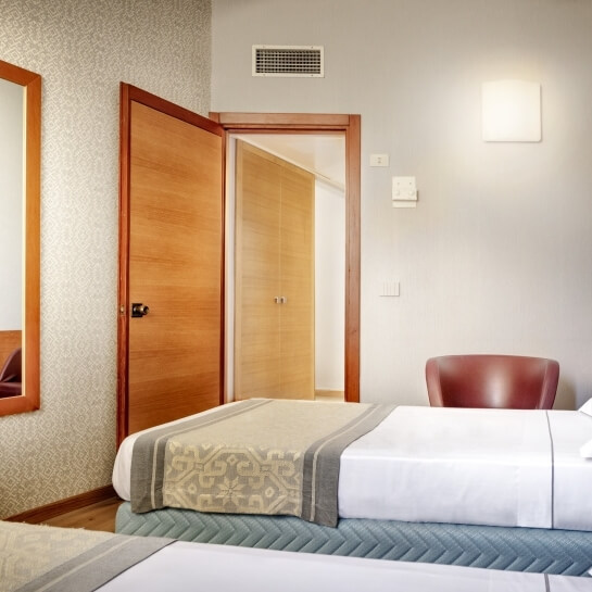 Camera d'albergo serena con letto matrimoniale, sedia salmonata, coperta geometrica e luce soffusa. Atmosfera rilassante.