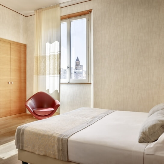 Camera serena con letto bianco, sedia rossa, armadio in legno e luce naturale che entra dalla finestra con vista su un grattacielo.