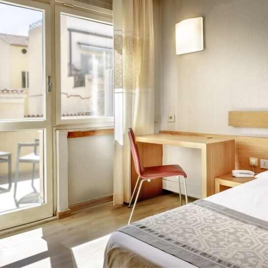 Camera d'hotel serena con decorazioni minimaliste, luce naturale, letto bianco e angolo scrivania con scaffale e telefono.