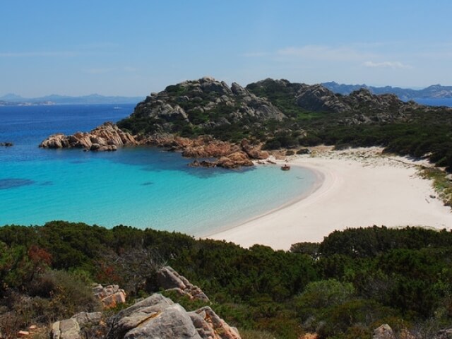 Una spiaggia serena con acqua blu, sabbia curva, rocce, vegetazione e un cielo azzurro con poche nuvole.