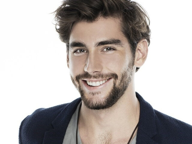 Uomo con capelli scuri e barba, indossa una camicia grigia e una giacca blu, sorridendo con uno sguardo pensieroso.