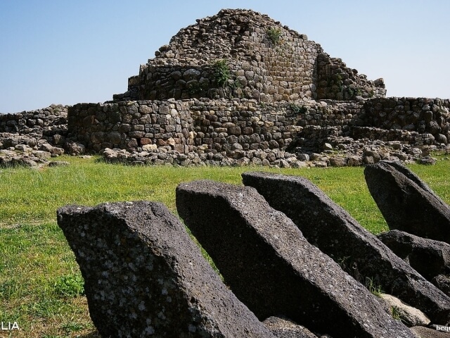 Campo erboso con grandi pietre scure in primo piano e una struttura simile a una piramide sullo sfondo, cielo blu.