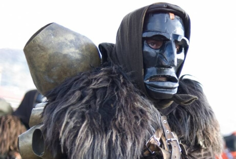 Persona in costume con maschera nera, colletto peloso e oggetto metallico sulla schiena, circondata da altri costumi.