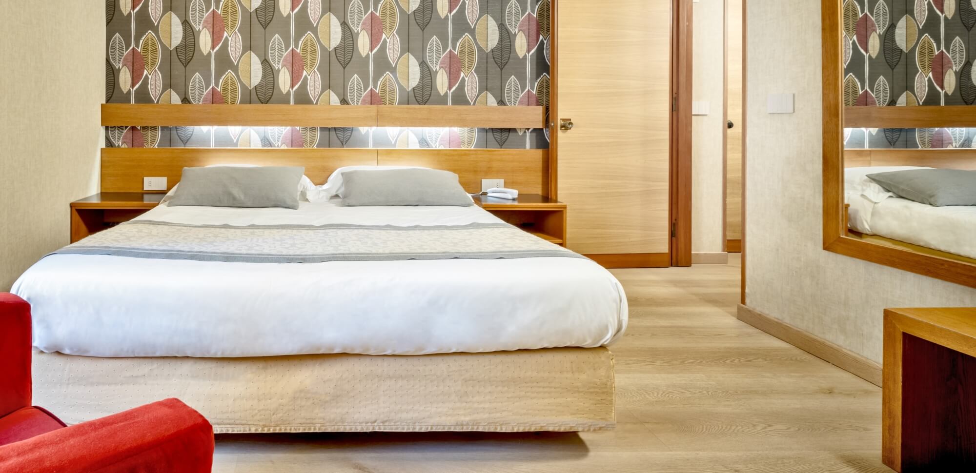 Camera d'hotel con letto bianco, testiera in legno e poltrona rossa.