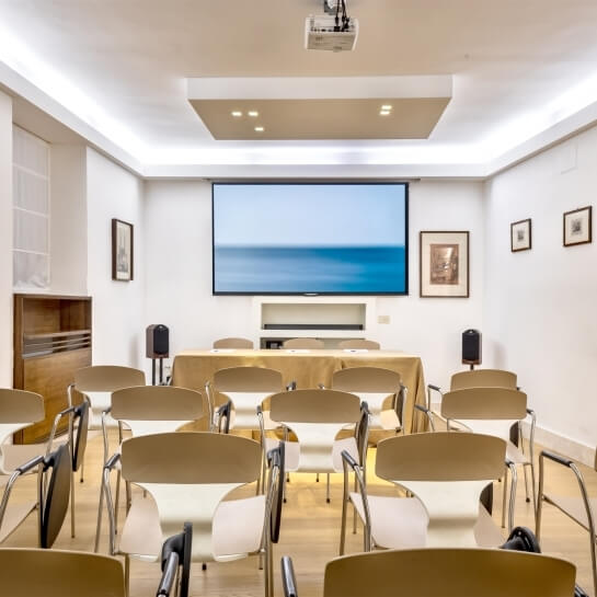 Sala riunioni con sedie, schermo proiettore e tavolo coperto.