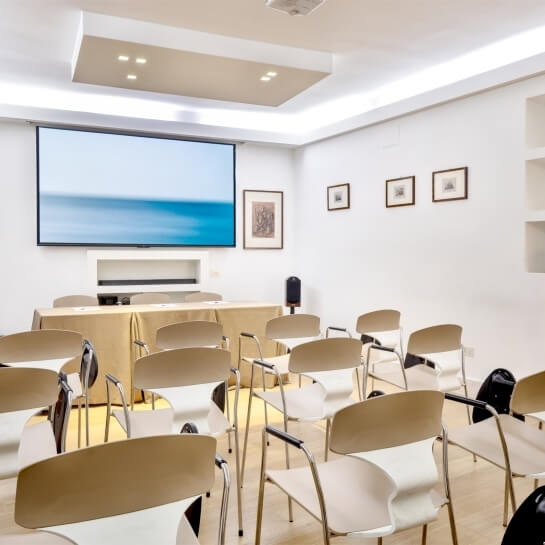 Sala conferenze con sedie, schermo, tavolo dorato e quadri.