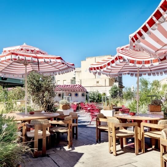 Outdoor dining area with wooden tables, striped umbrellas, and greenery.