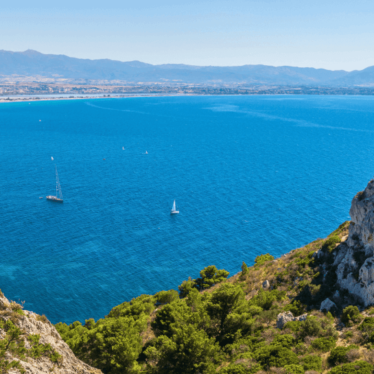 Una scena serena di un corpo d'acqua blu con velieri bianchi, una collina verde e montagne lontane sotto un cielo azzurro.