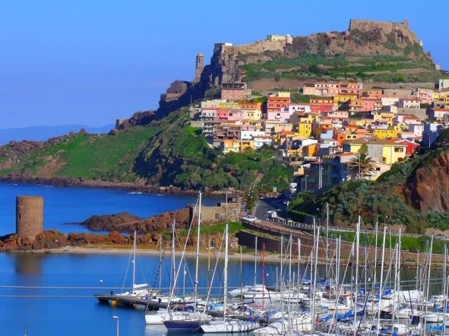 Una pittoresca città costiera colorata su una collina, con un marina affollato di barche a vela e una spiaggia sabbiosa.