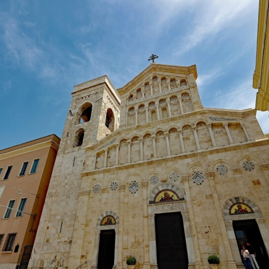 Chiesa con facciata in pietra chiara, torre campanaria, porte ad arco e finestre in vetro colorato, sotto un cielo blu.
