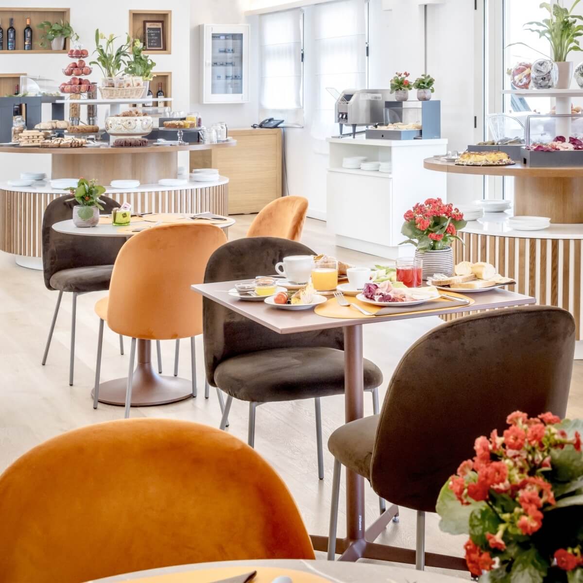 Bright breakfast area with white walls, wooden floors, tables with food, flowers, and curved bars, exuding warmth and hospitality.