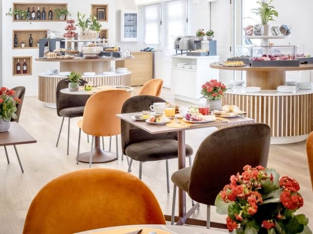 Sala colazioni moderna e luminosa con tavoli e sedie minimalisti, area di esposizione cibo e atmosfera elegante e accogliente.