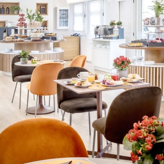 Area pranzo moderna e luminosa con tavoli rotondi, piatti di cibo, piante e un lungo bancone con cibi e bevande.