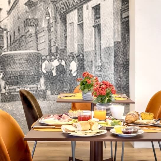 Tavolo lungo con una ricca colazione, piatti vari, piante e sedie moderne, sfondo di un murale grigio di edifici storici.