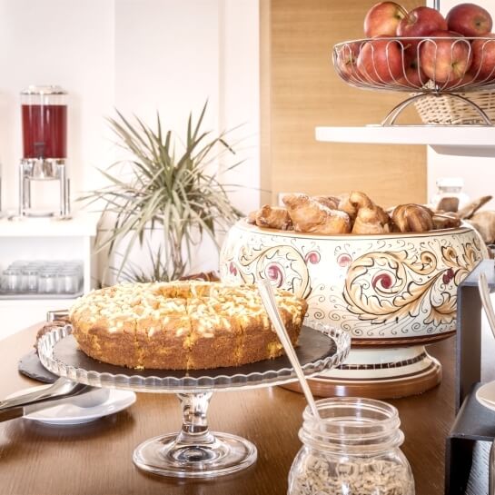 Un tavolo con una torta su un supporto, barattoli di ingredienti, una ciotola di pasticcini e una ciotola di frutta.