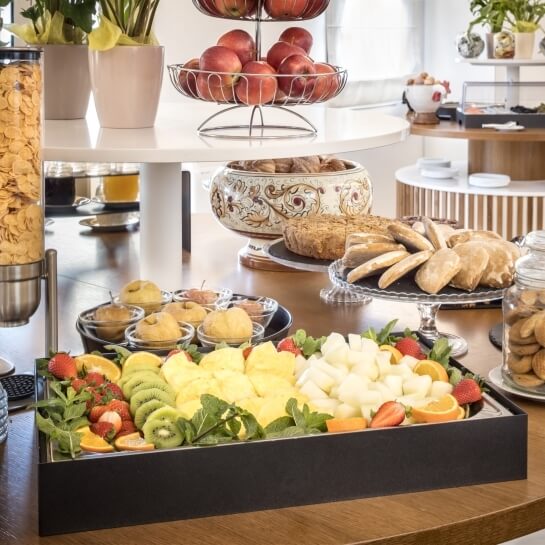 Tavolo buffet in legno chiaro con frutta, dolci, noci, mele, biscotti e piante, illuminato da finestre bianche.