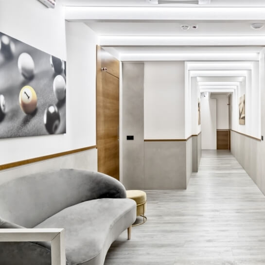 Un lungo corridoio bianco con porte, un divano grigio, un pouf giallo e un dipinto di palle da biliardo.