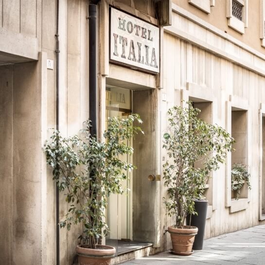 Ingresso dell'Hotel Italia con insegna, facciata in pietra chiara e piante in vaso, trasmette eleganza e raffinatezza.