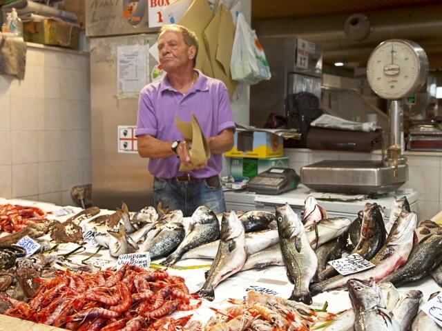 Uomo in polo viola dietro un banco di pesce, circondato da frutti di mare e con una busta di carta in mano.
