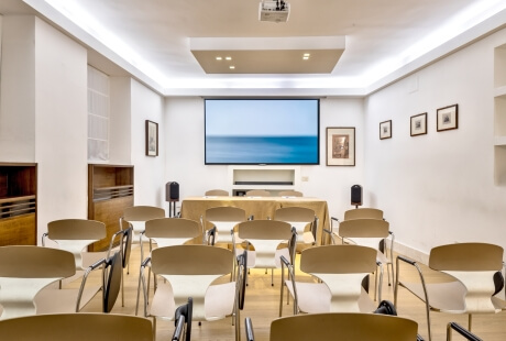Sala conferenze con sedie, schermo proiettore e tavolo decorato.