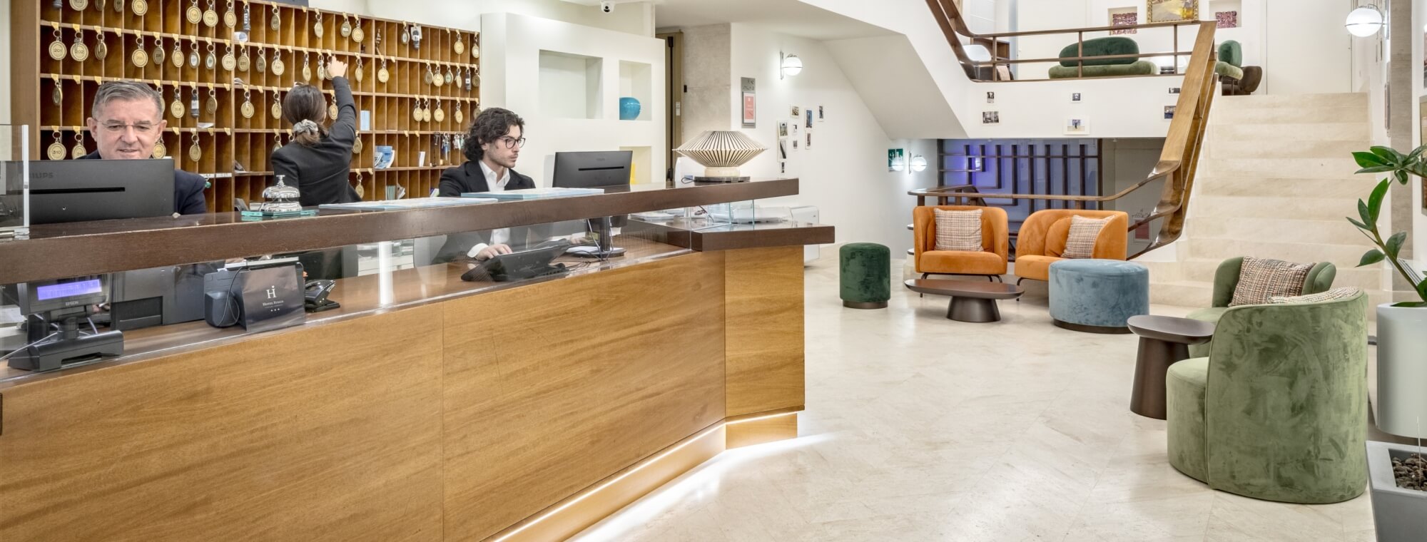 Reception area with staff, seating, and staircase in background. Area reception con personale, posti a sedere e scala sullo sfondo.