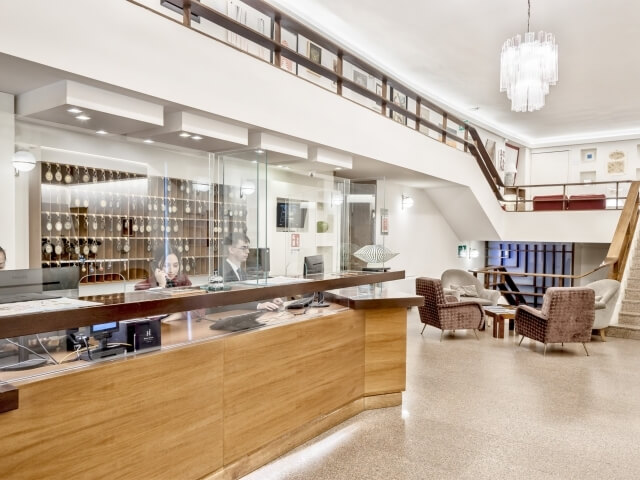 Hotel lobby with reception desk, two women working, bright decor, chandelier, staircase, and comfortable seating area.