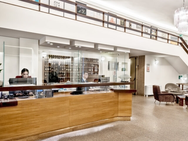 Modern office reception with a wooden desk, glass partition, lounge area, and a wall of glass panels displaying lights.