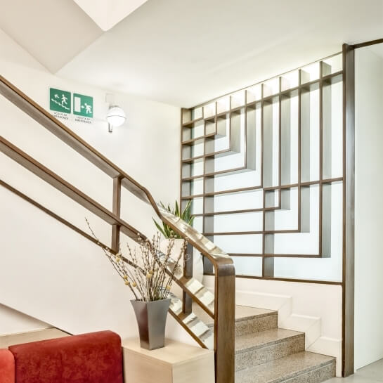 Scala in un edificio moderno con parete bianca, ringhiera metallica, piante in vaso e finestra decorativa.