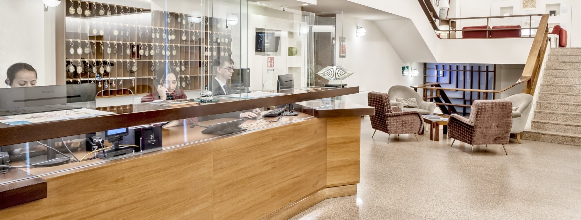 Lobby moderna con reception curva, personale, chiavi, TV e area salotto al piano superiore.