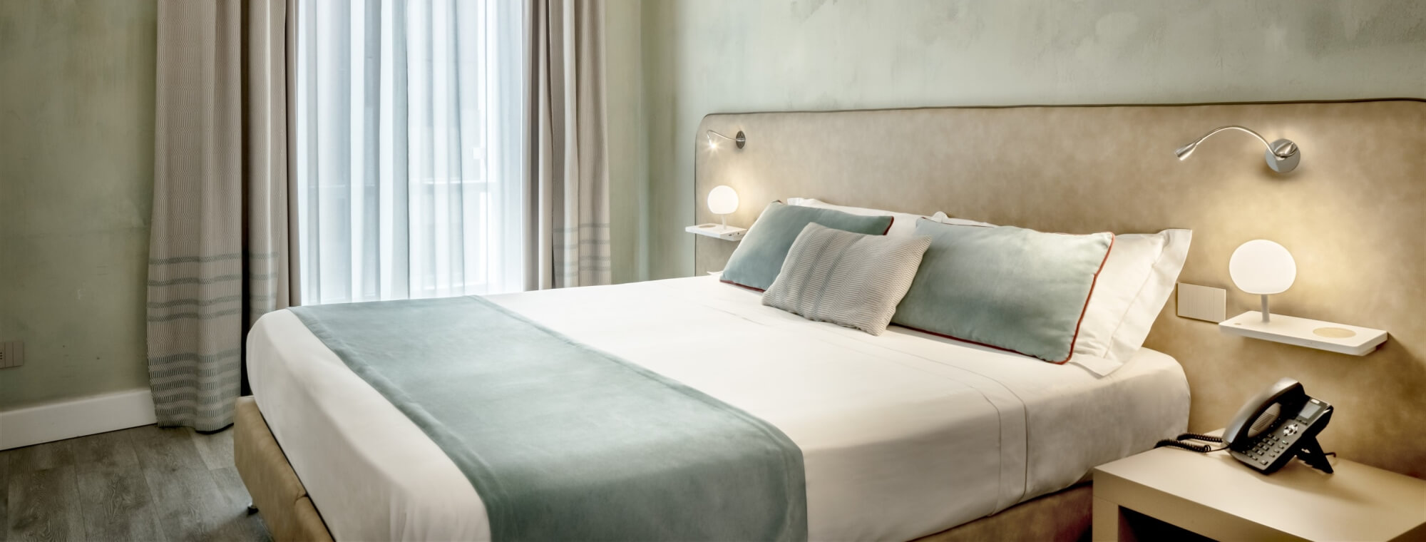 Hotel room with a large bed, blue and white pillows, soft lighting, and a calming green wall.