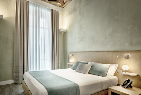 Camera da letto serena con letto bianco, comodino con lampada e telefono, e finestra con tende bianche parzialmente aperte.