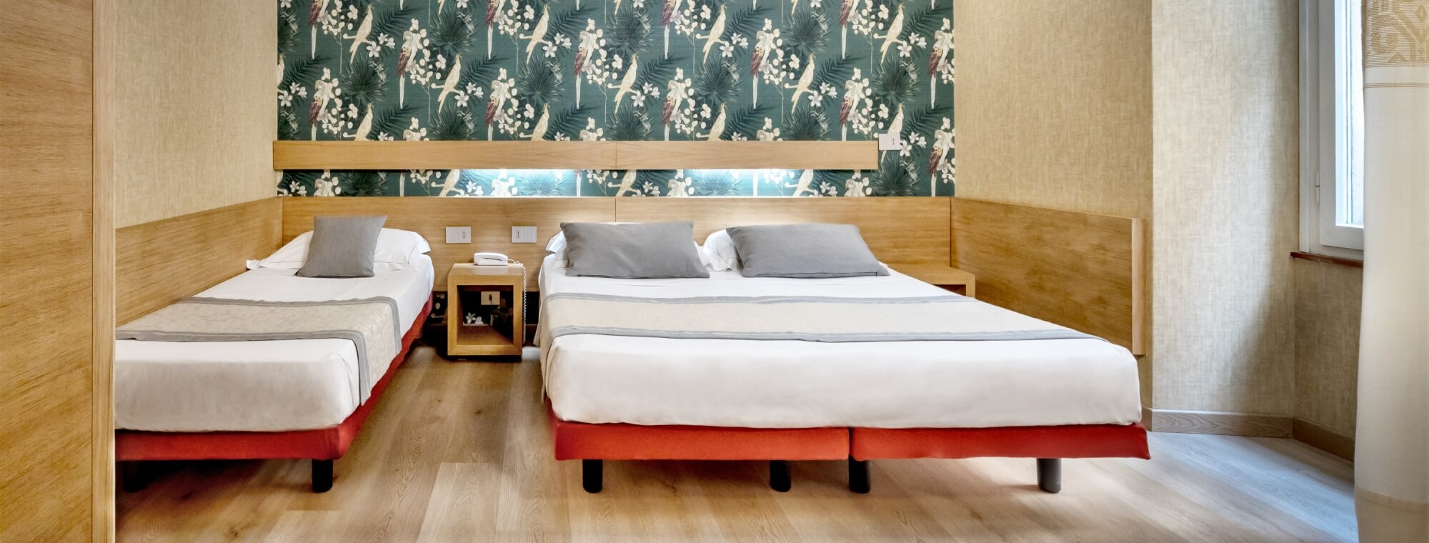 Cozy hotel room with two red-framed beds, tropical wallpaper, light wood floor, and a window letting in natural light.