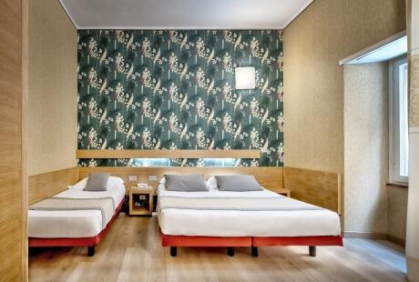 Camera d'hotel con due letti, comodino, finestra e carta da parati floreale verde e bianca dietro i letti.