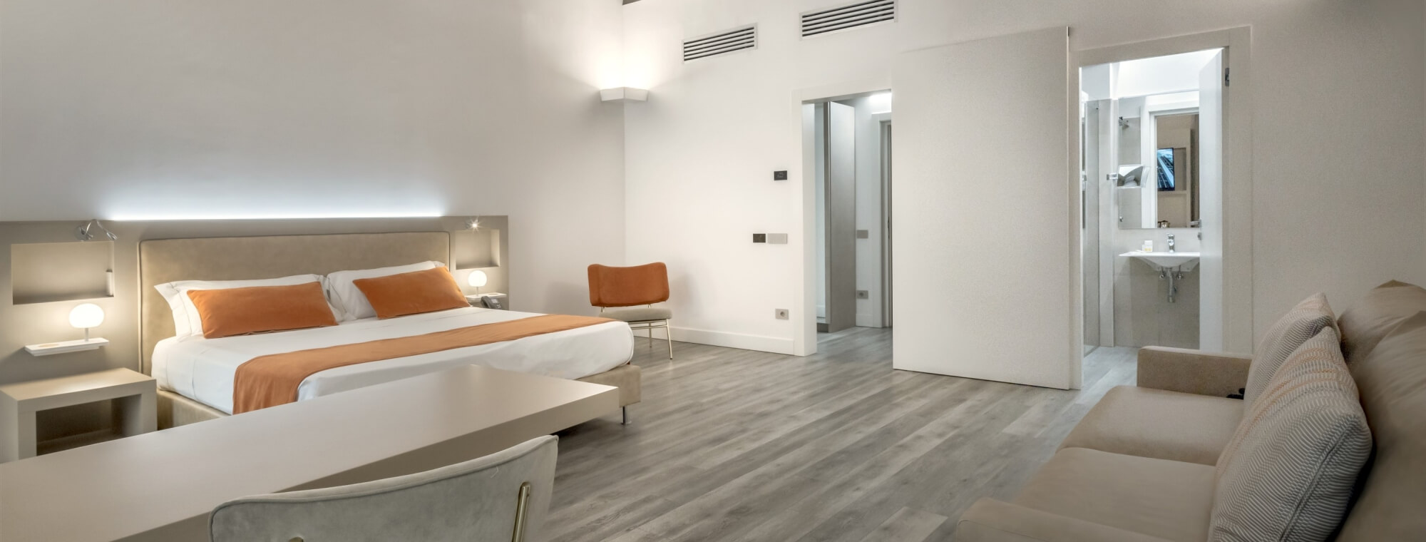 Modern hotel room with open living area, bed with orange throw, desk, couch, and natural light; kitchen and bathrooms visible.