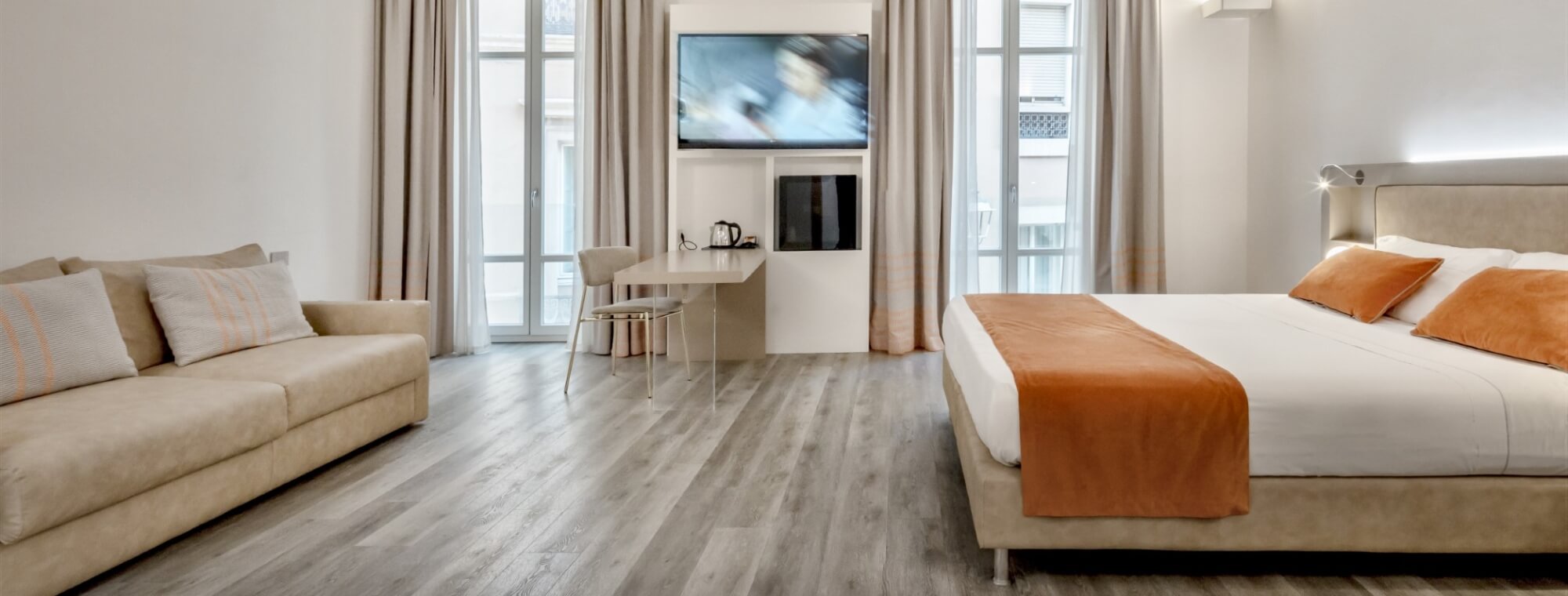 Modern hotel room with a king-size bed, beige sofa, kitchenette, and large windows, featuring a minimalist design.