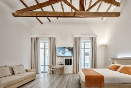 Spacious white room with living area and bedroom, featuring a beige couch, white bed, and rustic wooden beams.