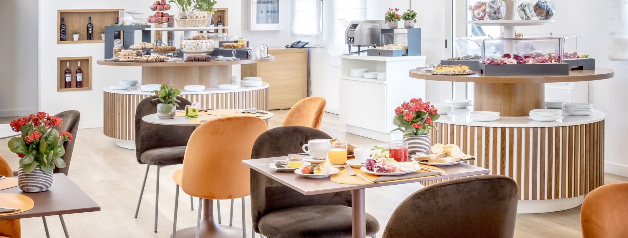 Bright cafeteria with buffet area, small tables with place settings, potted plants, and a wall of beverages in the background.