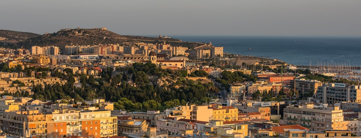 Vista panoramica di una città collinare al tramonto, con edifici vari e spazi verdi, e il mare sullo sfondo.