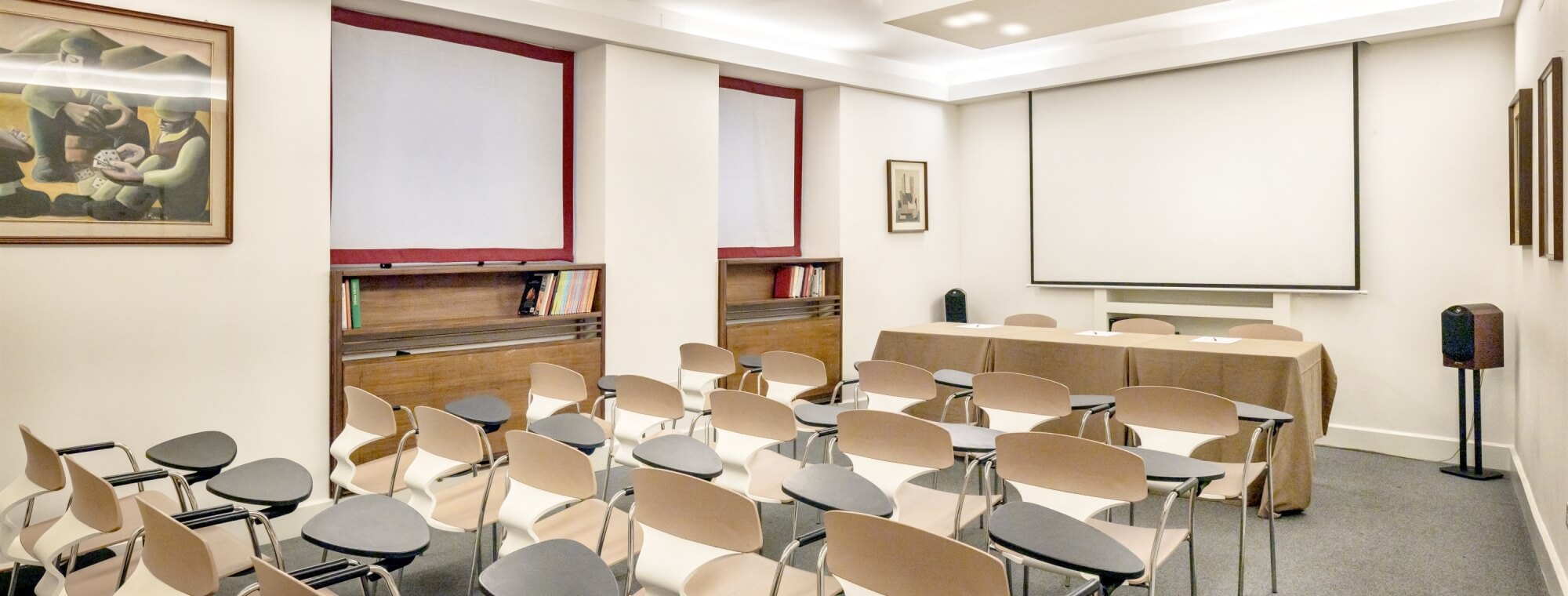 Conference room with projector screen, tan chairs, brown tables, bookshelves, and paintings, designed for presentations.