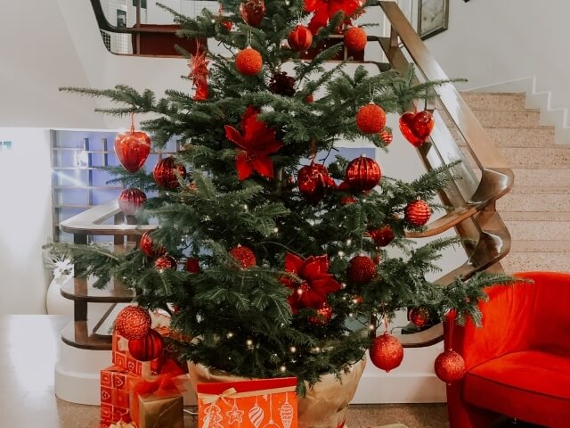 Albero di Natale decorato con ornamenti rossi, regali alla base, accanto a un divano rosso e una scala curva.