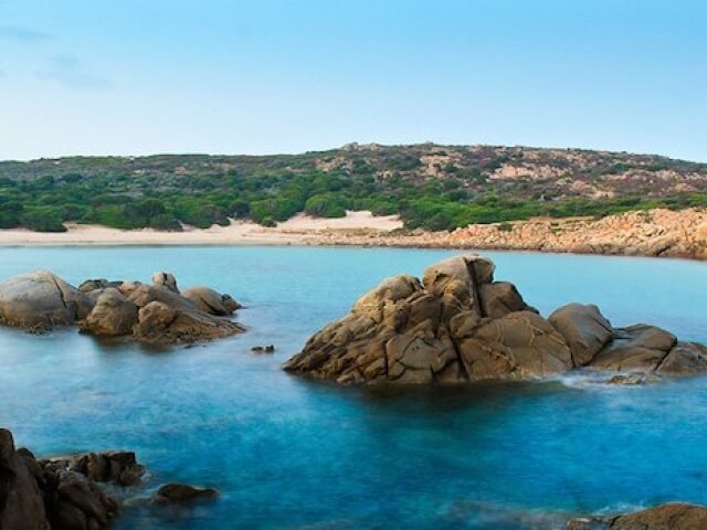 Una scena costiera serena con acqua blu, spiaggia sabbiosa, formazioni rocciose e cielo azzurro senza nuvole.