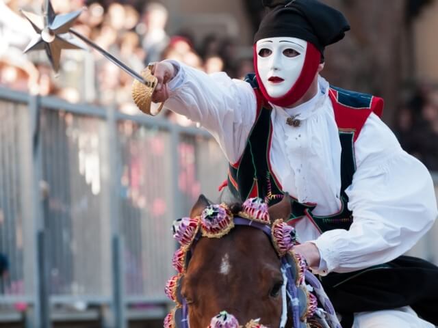 Una persona in abiti tradizionali cavalca un cavallo, impugnando una lancia decorativa, durante un evento festivo.