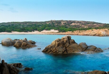 Una scena di spiaggia serena con grandi rocce, acqua calma e un paesaggio montano sullo sfondo sotto un cielo blu.