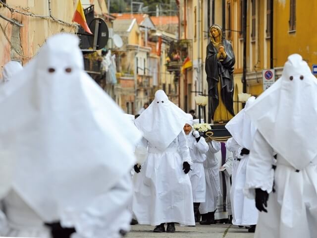 Gruppo di persone in tuniche bianche attorno a una statua dorata di una donna in abito scuro, in un'atmosfera religiosa.