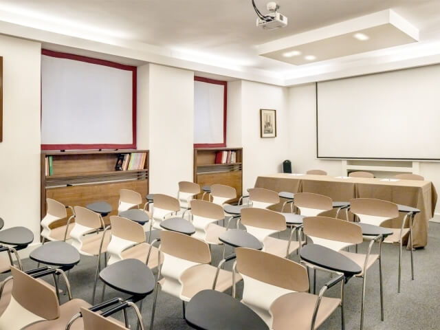 Aula con pareti bianche, proiettore, sedie con scrivania, tavolo, lavagne bianche e libreria, atmosfera professionale.