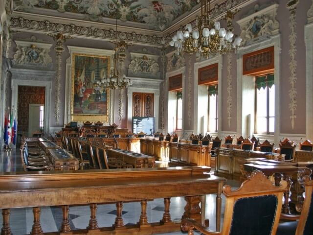 Una grande sala elegante con tavoli e sedie disposti in fila, illuminata da luce naturale e decorata con un lampadario.