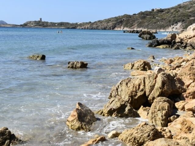 Una tranquilla costa rocciosa con acqua cristallina, colline verdi e persone che nuotano, esprimendo serenità e bellezza naturale.