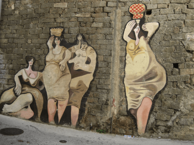 Mural di quattro donne in abiti bianchi su un muro di mattoni, in uno stile simile a Picasso, con colori tenui.